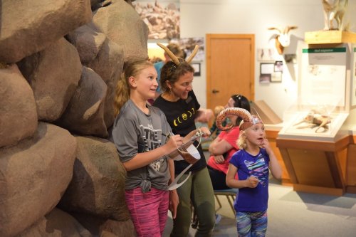 Three people, including two children, wearing animal-themed headbands, engage in an interactive exhibit in a museum.