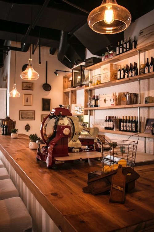 A cozy café interior featuring a wooden bar, vintage coffee machine, and shelves lined with wine bottles and decor.