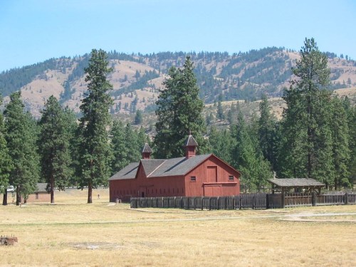 A red barn surrounded by tall pine trees and mountains under a clear blue sky.