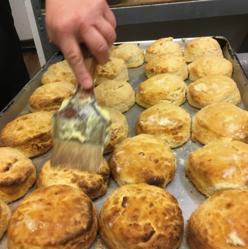 A hand brushes melted butter onto freshly baked scones arranged on a baking tray.