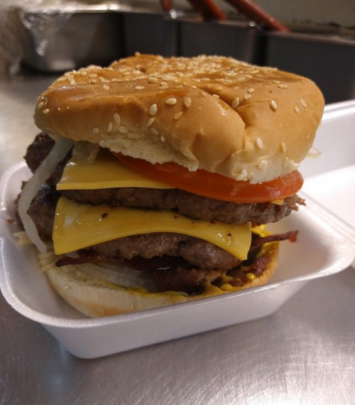A stacked burger with two beef patties, cheddar cheese, tomato, and onions on a sesame seed bun in a takeout container.
