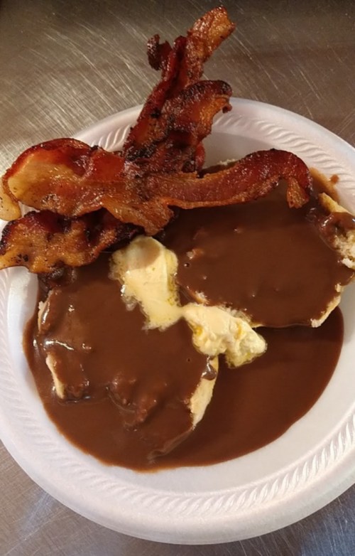A plate with two biscuits topped with brown gravy and crispy bacon strips on the side.
