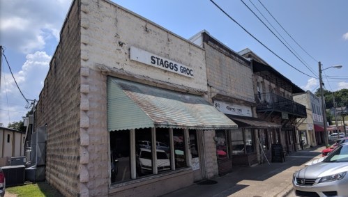 Exterior of a small grocery store named "Staggs Groc," with a corrugated awning and nearby buildings on a sunny day.