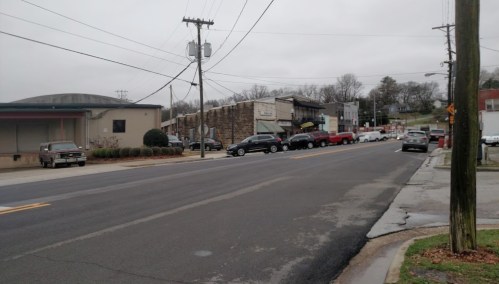 A street scene with parked cars, buildings, and power lines under a cloudy sky.