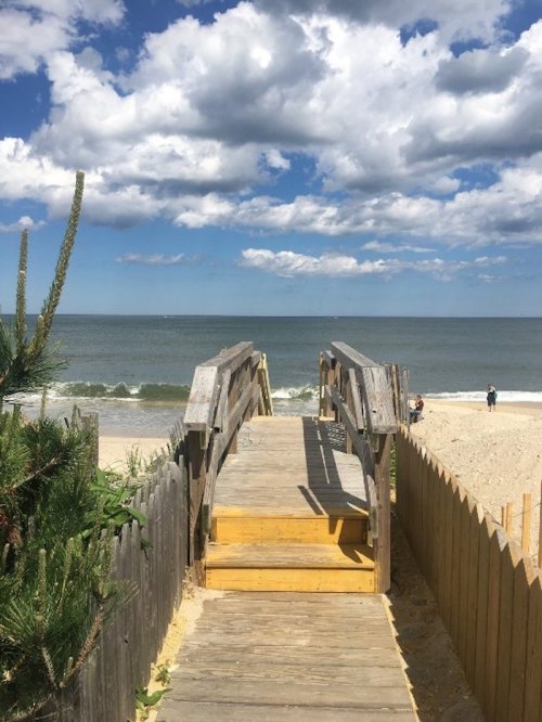 A wooden walkway leads to a sandy beach under a blue sky with fluffy clouds and gentle waves.