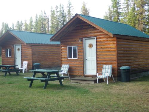 Two wooden cabins with green roofs, surrounded by trees, featuring picnic tables and chairs in a grassy area.