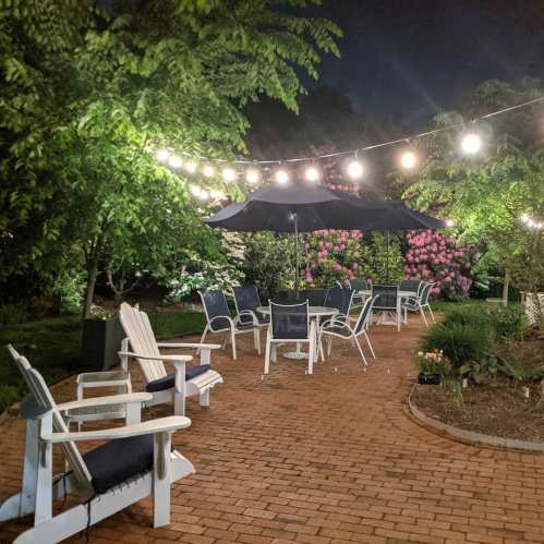 A cozy outdoor patio at night, featuring string lights, an umbrella, and seating surrounded by lush greenery and flowers.