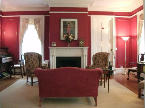Cozy living room with red walls, floral armchairs, a fireplace, and a portrait above a mantel. Soft lighting enhances the ambiance.