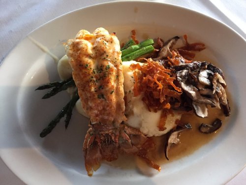A plated dish featuring lobster, mashed potatoes, asparagus, and crispy onions, garnished with herbs.