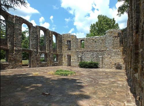 Ruins of a stone building with arched windows, surrounded by grass and trees under a partly cloudy sky.