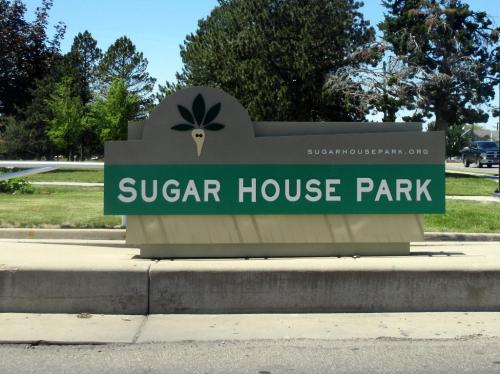 Sign for Sugar House Park, featuring a carrot logo and website URL, set against a green park backdrop.