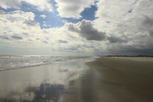 A serene beach scene with gentle waves, cloudy skies, and reflections on the wet sand.