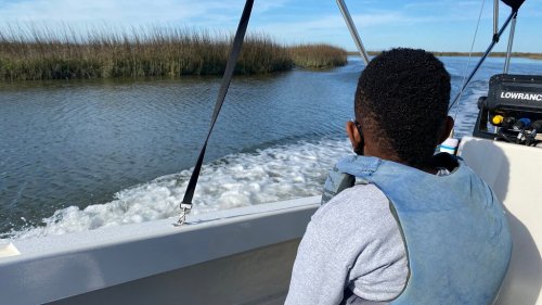 A child in a life jacket sits on a boat, looking out at a calm waterway surrounded by tall grass.