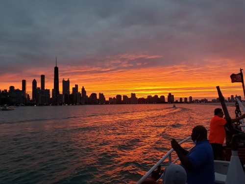 A vibrant sunset over a city skyline, with silhouettes of buildings and people on a boat in the foreground.