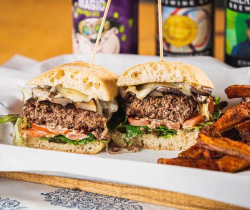A delicious burger with melted cheese, lettuce, and tomato, served with sweet potato fries and drinks in the background.