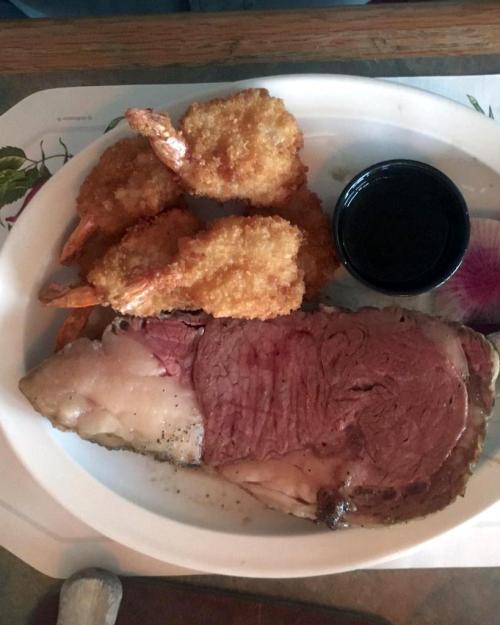 A plate featuring a slice of prime rib, two breaded shrimp, and a small cup of dipping sauce.