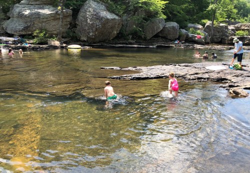 Two children play in a shallow river surrounded by rocks and greenery, with other people enjoying the water nearby.