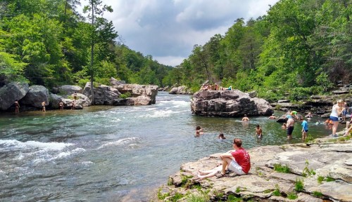 People enjoying a sunny day by a river, swimming and relaxing among rocks and lush greenery.