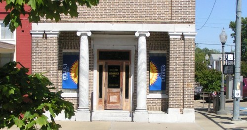 A brick building with columns, featuring banners and a wooden door, set in a sunny urban environment.