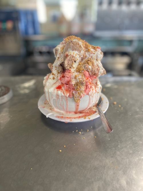 A dessert bowl topped with whipped cream, fruit, and a drizzle of syrup, sitting on a plate with a spoon beside it.