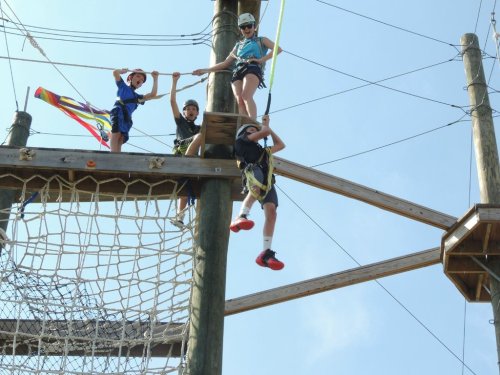Four children participate in a high ropes course, climbing and hanging on wooden structures against a blue sky.