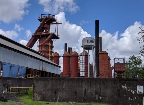 Industrial site featuring red silos, a water tower labeled "SLOSS," and a blue sky with scattered clouds.