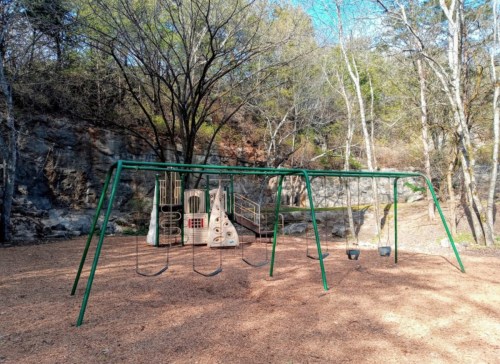 A playground with swings and a climbing structure, surrounded by trees and rocky terrain.