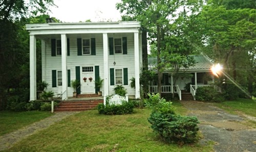 A charming white two-story house with green shutters, surrounded by trees and a grassy yard.