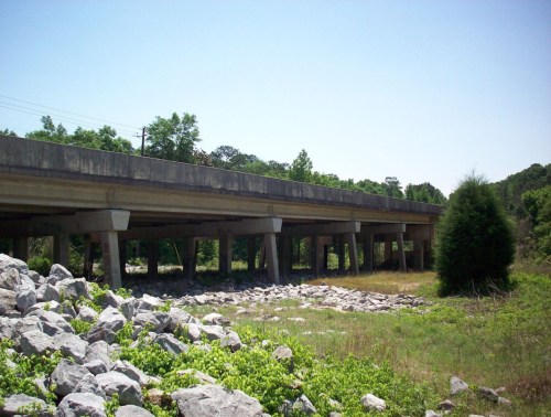 A concrete overpass supported by pillars, surrounded by greenery and large rocks under a clear blue sky.