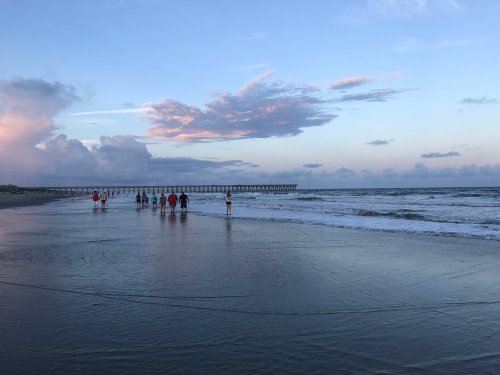 People walking along a beach at sunset, with gentle waves and a pier in the background under a colorful sky.