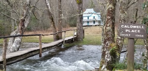 A wooden bridge over a flowing stream leads to a historic blue house, with a sign reading "Caldwell Place, Built 1903."