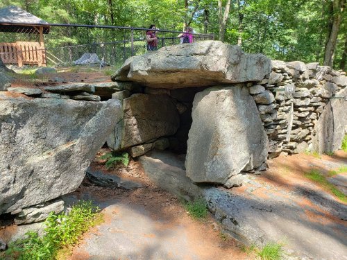 A stone structure resembling a dolmen, surrounded by trees and a pathway, with people visible in the background.