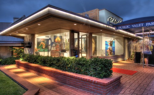 A modern art gallery entrance with glass walls, illuminated at night, featuring a red carpet and lush greenery.