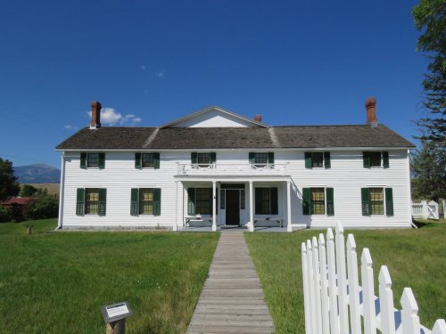 A large white historic house with green shutters, surrounded by grass and a white picket fence under a clear blue sky.