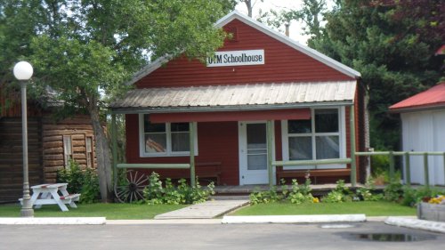 A red schoolhouse with a sign reading "MUM Schoolhouse," surrounded by greenery and a white picnic table.