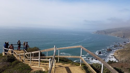 A scenic coastal view with people standing on a path overlooking the ocean and cliffs under a cloudy sky.