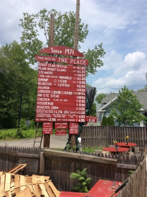 A weathered menu board listing seafood and other dishes with prices, set in a rustic outdoor area.