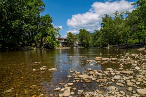 A serene river scene with clear water, smooth stones, and lush green trees under a blue sky with fluffy clouds.