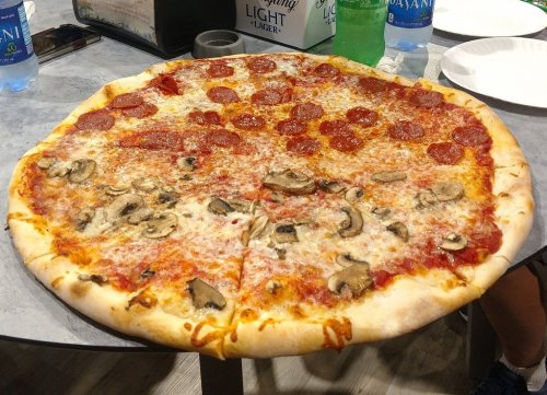 A large pizza topped with pepperoni and mushrooms, sitting on a table with drinks in the background.