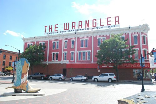 Historic building with "The Wrangler" sign, featuring a large cowboy boot sculpture in front. Bright blue sky above.