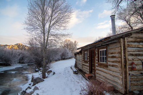 A rustic log cabin by a snowy riverbank, surrounded by frosty trees under a clear blue sky.