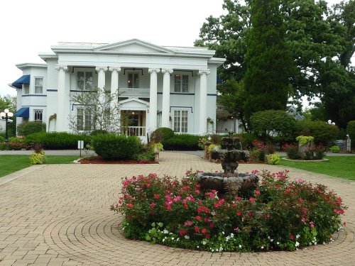 A grand white mansion with columns, surrounded by lush gardens and a flowerbed featuring a central fountain.