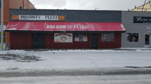 A snow-covered street scene featuring Broadway Pizza with a bright pink awning and a neighboring shop.