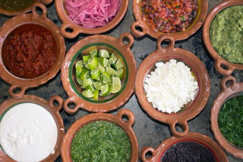A variety of colorful salsas and toppings in clay bowls, including limes, cheese, guacamole, and pickled onions.