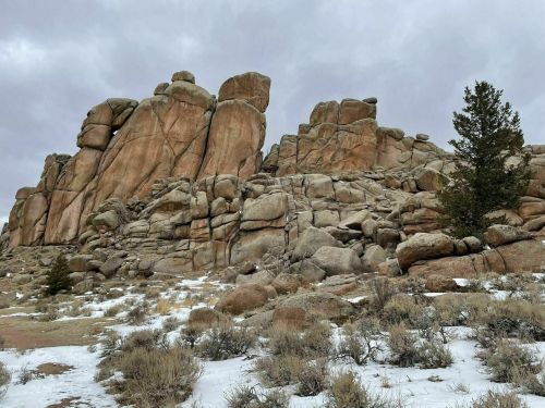 Rocky outcrop with large boulders, sparse vegetation, and a cloudy sky in the background. Snow patches on the ground.