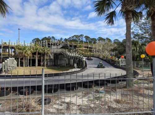 Go-karts racing on a winding track surrounded by palm trees and a wooden structure, with orange balloons in the background.