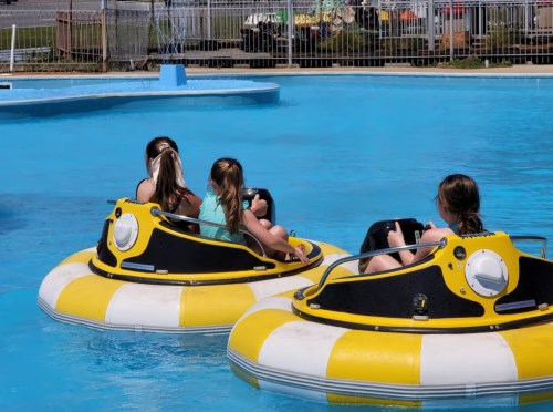 Three children in yellow and black bumper boats enjoying a sunny day in a blue pool.