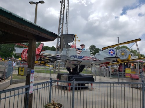 A child sits in a colorful airplane ride at an amusement park, surrounded by a fenced area and cloudy skies.