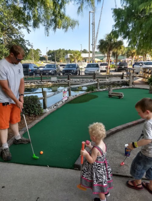 A man plays mini-golf while two young children watch, one holding a toy and the other in a dress.