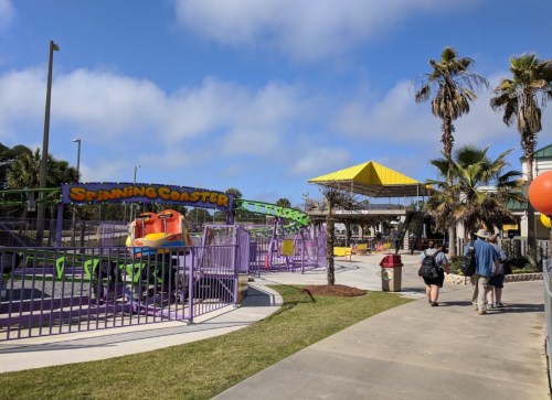 A colorful amusement park area featuring a spinning coaster, palm trees, and visitors walking along a paved path.